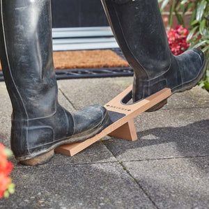 Black rubber wellington boots being removed using a wooden boot jack on a paved garden path with red flowers and a doormat in the background