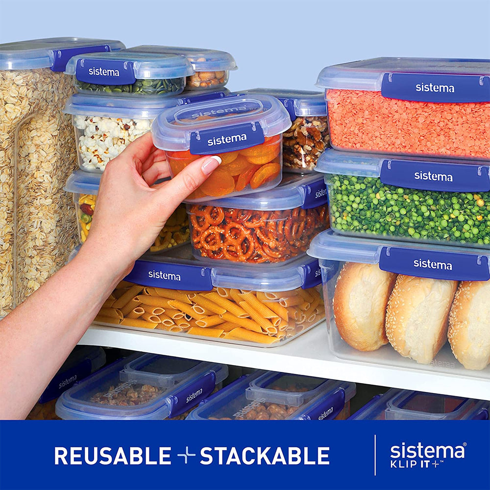 Hand holding a blue-lidded Sistema Klip It Plus clear plastic storage container filled with orange dried apricots, surrounded by stacked containers with various dry foods including oats, pasta, lentils, peas and bread rolls on white shelving, demonstrating reusable and stackable storage organization