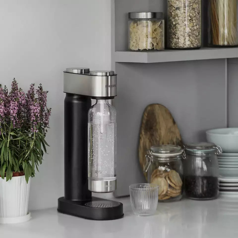 Philips GoZero black and stainless steel soda water maker on kitchen counter with carbonated water bottle, glass, flowers, storage jars and bread basket in background