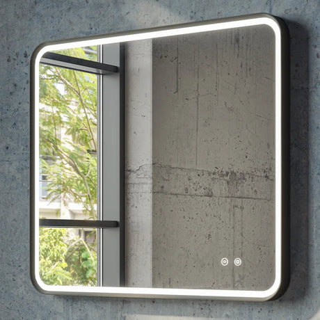 Modern LED bathroom mirror with rounded corners and gunmetal grey frame mounted on concrete wall, featuring illuminated edges, touch controls, and reflection of window with green foliage