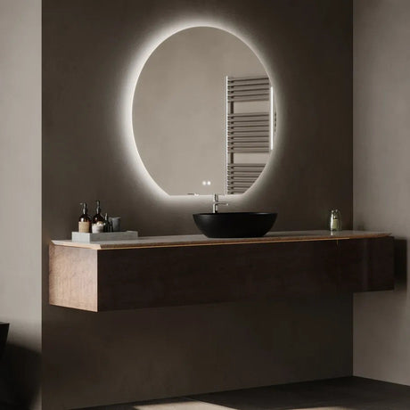 Modern bathroom vanity with oval LED-lit mirror, black ceramic sink, dark wood floating cabinet, chrome towel rail, and warm white wall lighting in contemporary minimalist style