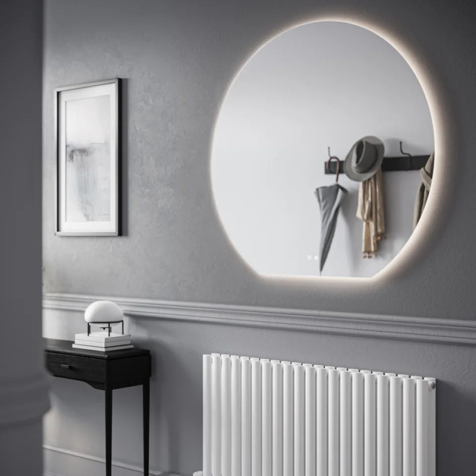 Modern illuminated oval LED bathroom mirror with warm backlight mounted on grey wall above white vertical radiator, styled with black side table, small decorative items, and hat rack with accessories