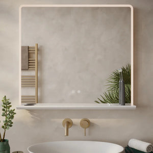 Modern minimalist bathroom mirror with integrated LED lighting and white frame mounted above a white oval sink with brass wall-mounted faucet, displaying a reflective surface showing a wooden towel rack, green potted plant and bathroom accessories on a light neutral wall