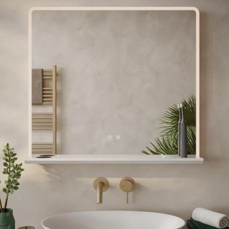 Modern minimalist bathroom mirror with integrated LED lighting and white frame mounted above a white oval sink with brass wall-mounted faucet, displaying a reflective surface showing a wooden towel rack, green potted plant and bathroom accessories on a light neutral wall