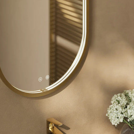 Modern Aqualla Solaris Pill LED bathroom mirror with brushed brass frame and integrated LED lighting, featuring touch controls and demister pad, mounted above beige wall with white flowers and gold towel rail