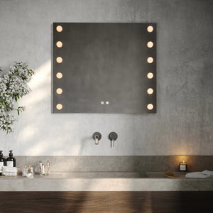 Modern bathroom vanity setup featuring Aqualla Studio LED mirror with warm-lit bulbs, concrete grey walls, white countertop, dark wood cabinetry, chrome wall-mounted faucet, and decorative accessories including potted plant and bathroom products