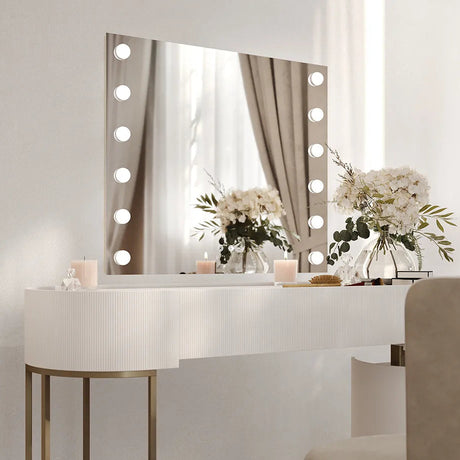 Modern bathroom vanity setup featuring a white ribbed curved desk with brass legs, illuminated LED mirror with 20 warm white bulbs around the frame, white flowers in glass vases, candles, and brushes on the countertop, with beige curtains visible in the mirror reflection, contemporary minimalist aesthetic