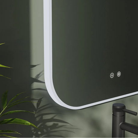 Modern arched LED bathroom mirror with white frame and dark green wall background, featuring touch controls and ambient lighting, displayed with black faucet and green plant foliage