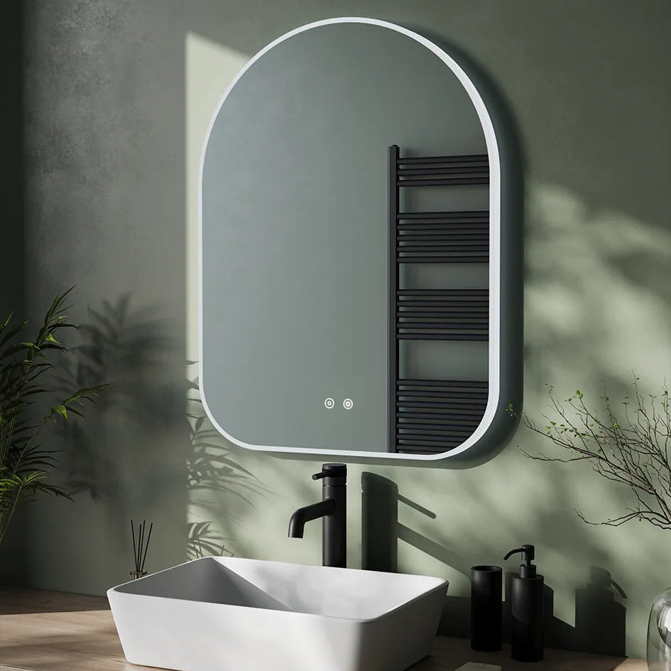 Modern bathroom vanity setup featuring an arched LED mirror with white frame above a white rectangular vessel sink, black matte faucet, black towel radiator, and black soap dispensers against a textured sage green wall with natural sunlight and decorative plants