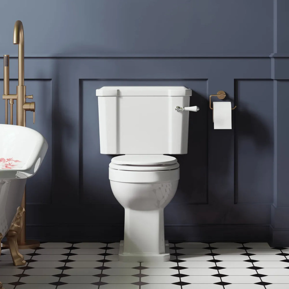 White ceramic close coupled toilet with dual flush cistern in a modern bathroom featuring blue-grey panelled walls, brass fittings, black and white geometric floor tiles, and matching brass toilet paper holder