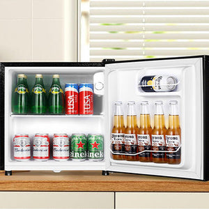 Black compact table top refrigerator with open door displaying various beverages including green bottles, red and pink cans, and brown bottles arranged on white shelves, positioned on wooden surface with white blinds visible in background