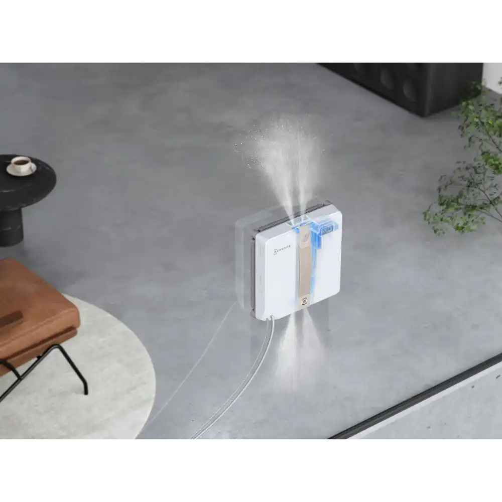 Ecovacs Winbot Mini white and beige robot window vacuum in action spraying water mist on an outdoor patio window with concrete flooring and garden furniture visible