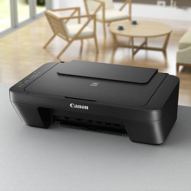 Canon PIXMA MG2551S all-in-one printer in black displayed on a white desk with wooden chairs and bright interior visible in background