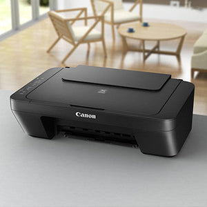 Canon PIXMA MG2551S all-in-one printer in black displayed on a white desk with wooden chairs and bright interior visible in background