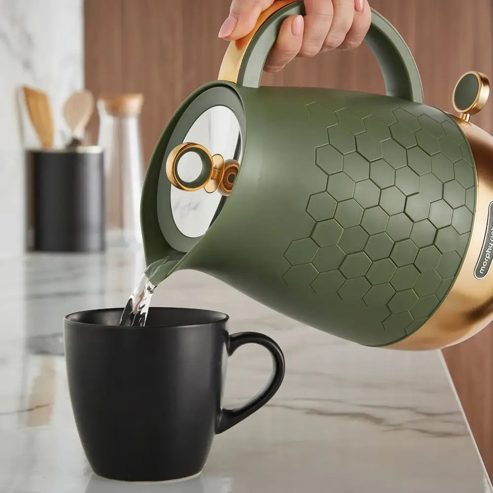 Morphy Richards Cassini jug kettle in olivine green with gold accents pouring hot water into a black mug, featuring a honeycomb textured body and modern design on a marble countertop