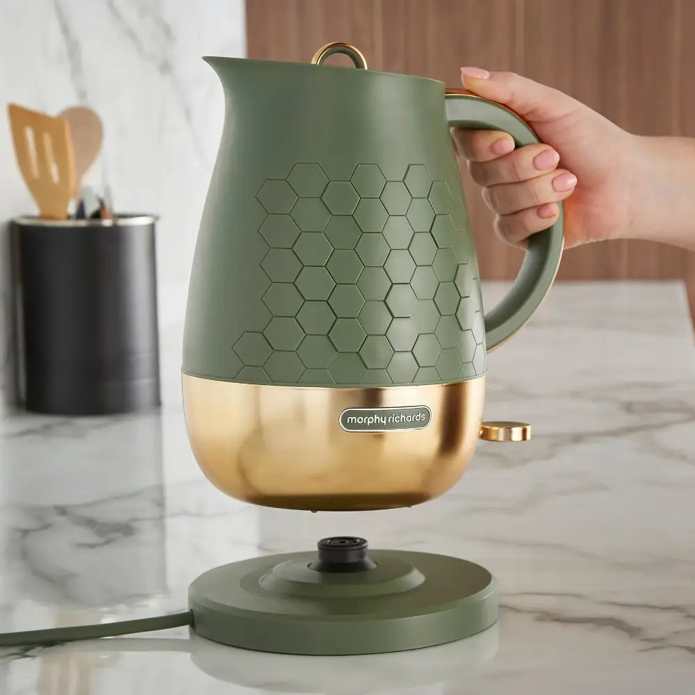 Morphy Richards Cassini electric jug kettle in sage green with gold base, featuring honeycomb textured design, shown being held with matching base station on marble countertop next to kitchen utensil holder