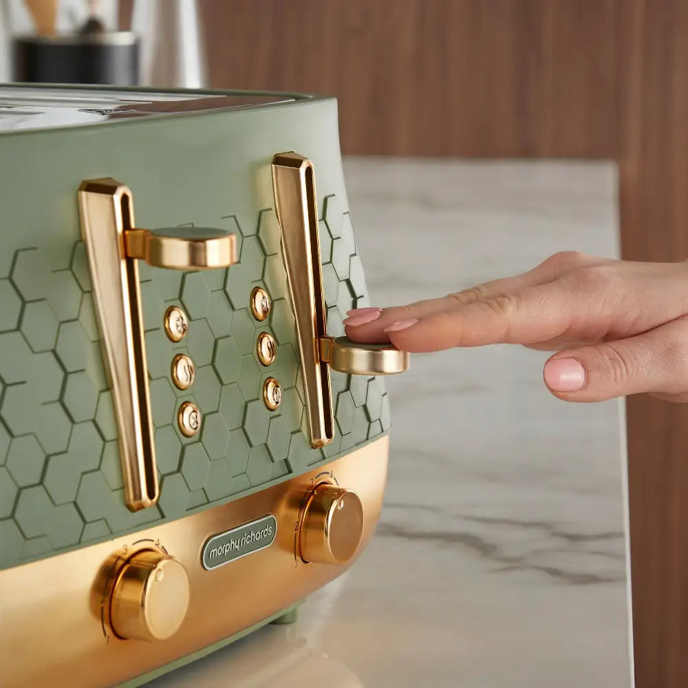 Person demonstrating the lever mechanism on a Morphy Richards Cassini 4-slice toaster with sage green hexagonal tile pattern body, rose gold metal accents, and control dials on the base
