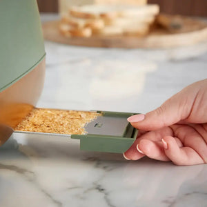 Hand holding a green measuring scoop filled with brown sugar or granulated ingredient over a marble countertop with kitchen appliances blurred in the background