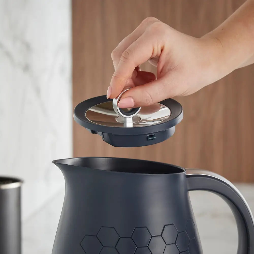 Hand lifting the stainless steel lid off a dark grey Morphy Richards Cassini jug kettle with hexagonal pattern design, showing the interior spout against a warm brown and white interior background