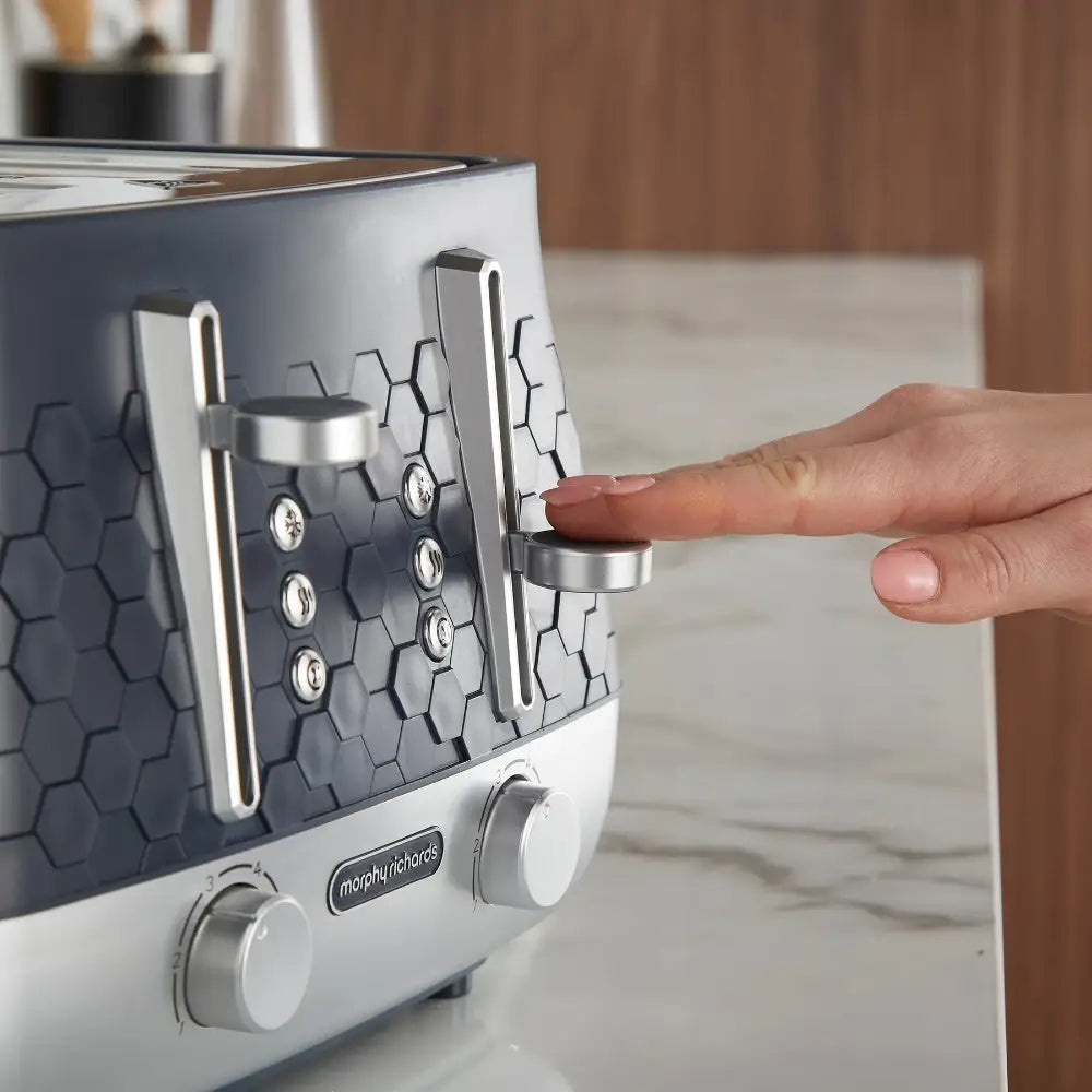 Hand pressing the lever on a Morphy Richards Cassini 4 slice toaster in white and dark gray with hexagonal pattern design, featuring chrome accents and control dials on a marble countertop