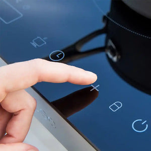 Close-up of a hand touching the blue digital display panel of a Cecotec double induction hob, showing illuminated touch controls including power button, temperature settings, and timer icons with black bezels and sleek modern design