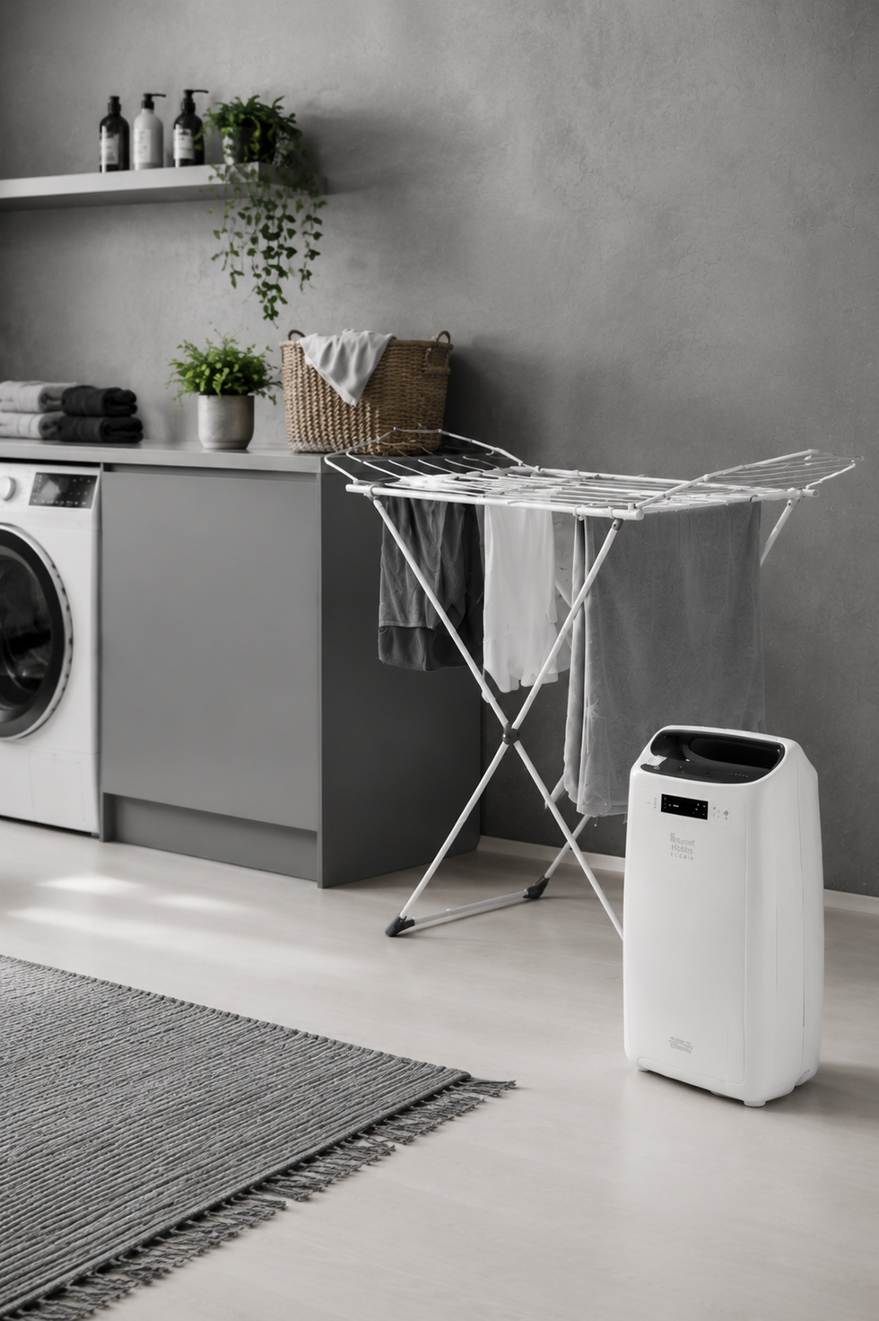 A white portable air conditioner or heater placed on a light floor in a modern laundry room, next to a metal folding clothes rack and a gray washing machine.