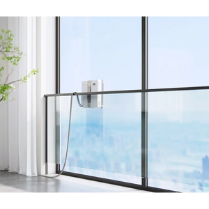 Ecovacs Winbot Mini beige robot window vacuum mounted on modern floor-to-ceiling window with black frame, cleaning glass pane with city skyline view, white curtains and green plant visible to the left