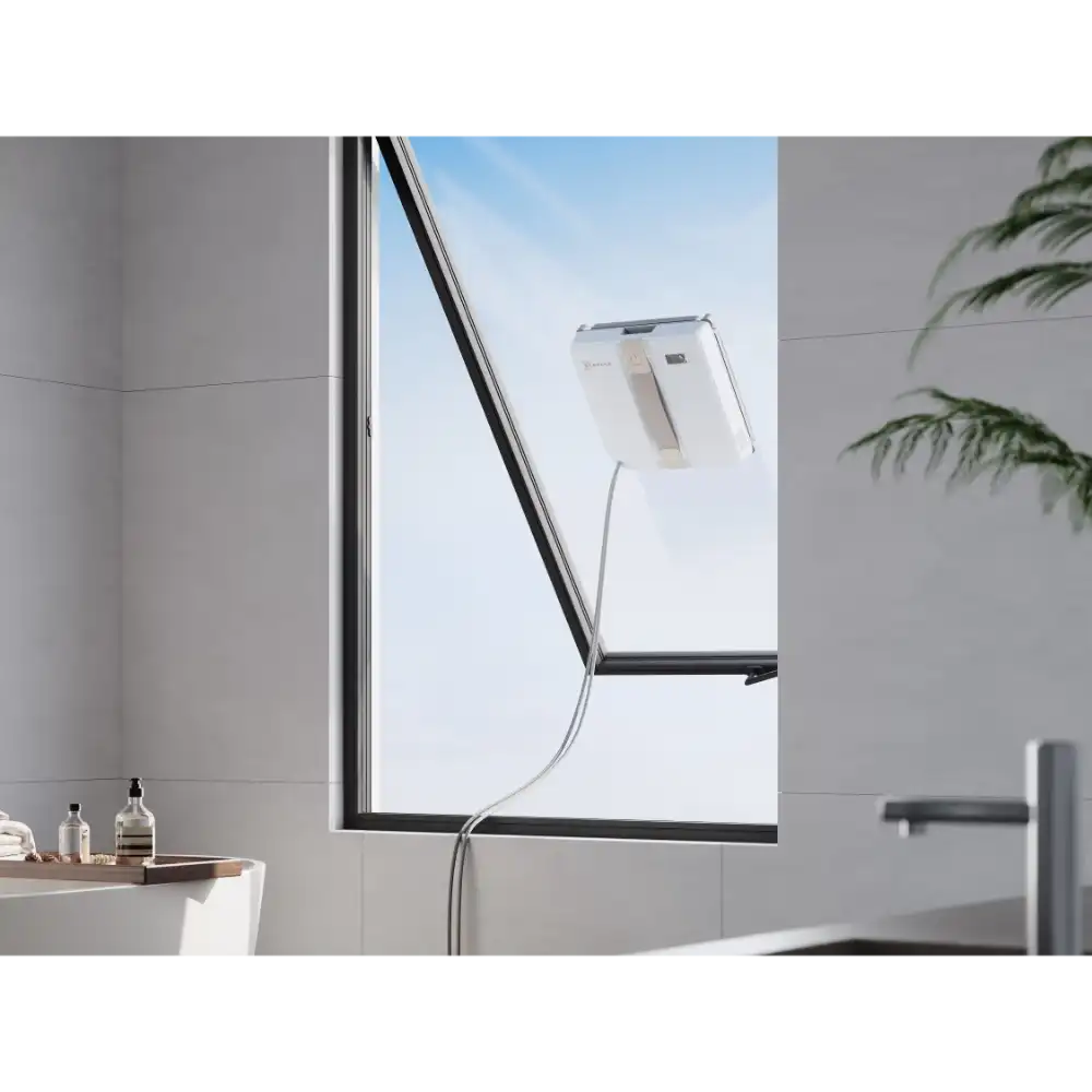 Ecovacs Winbot Mini beige robot window vacuum cleaner attached to exterior window glass with power cable, shown in modern bathroom setting with grey tiled walls and potted plant