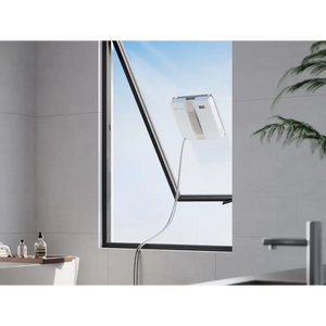 Ecovacs Winbot Mini beige robot window vacuum cleaner attached to exterior window glass with power cable, shown in modern bathroom setting with grey tiled walls and potted plant
