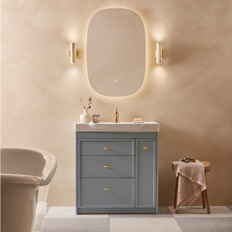 Modern bathroom vanity unit with matt carbon grey finish, white basin top, three drawers with brass handles, matching oval illuminated mirror with gold wall sconces, styled with neutral beige walls, decorative accessories and wooden stool