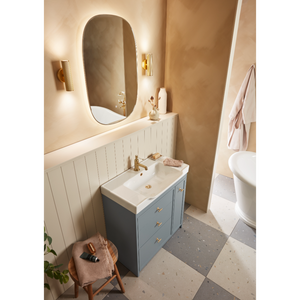 Modern bathroom vanity unit featuring a Roper Rhodes Concierge basin in matt Coniston blue with white ceramic sink, brass tap and handles, paired with an oval illuminated mirror, white shiplap panelling, warm beige walls, brass wall sconces, terrazzo flooring, and coordinating accessories in a contemporary spa-like setting