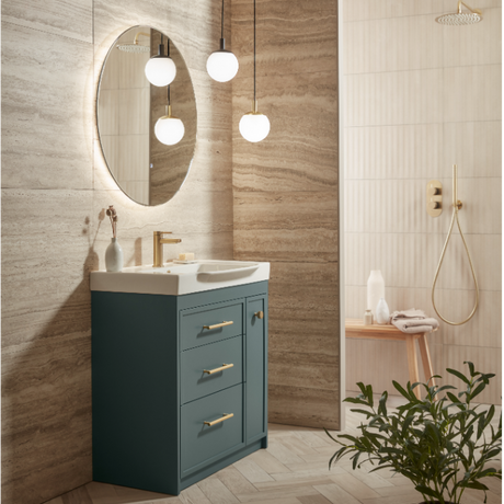 Modern bathroom vanity unit with matt Juniper Green cabinetry, white ceramic basin, brushed brass handles, oval illuminated mirror, pendant lighting, and travertine tile walls in a contemporary spa-like setting
