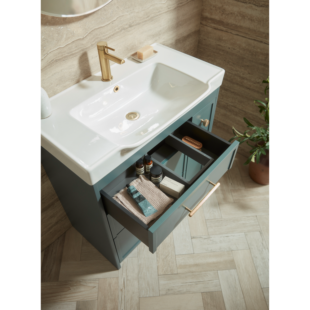 Roper Rhodes Concierge vanity unit with white ceramic basin, matt coniston blue cabinet, brass tap, open drawer displaying toiletries and towels, herringbone wood flooring, stone tile walls, modern bathroom setting