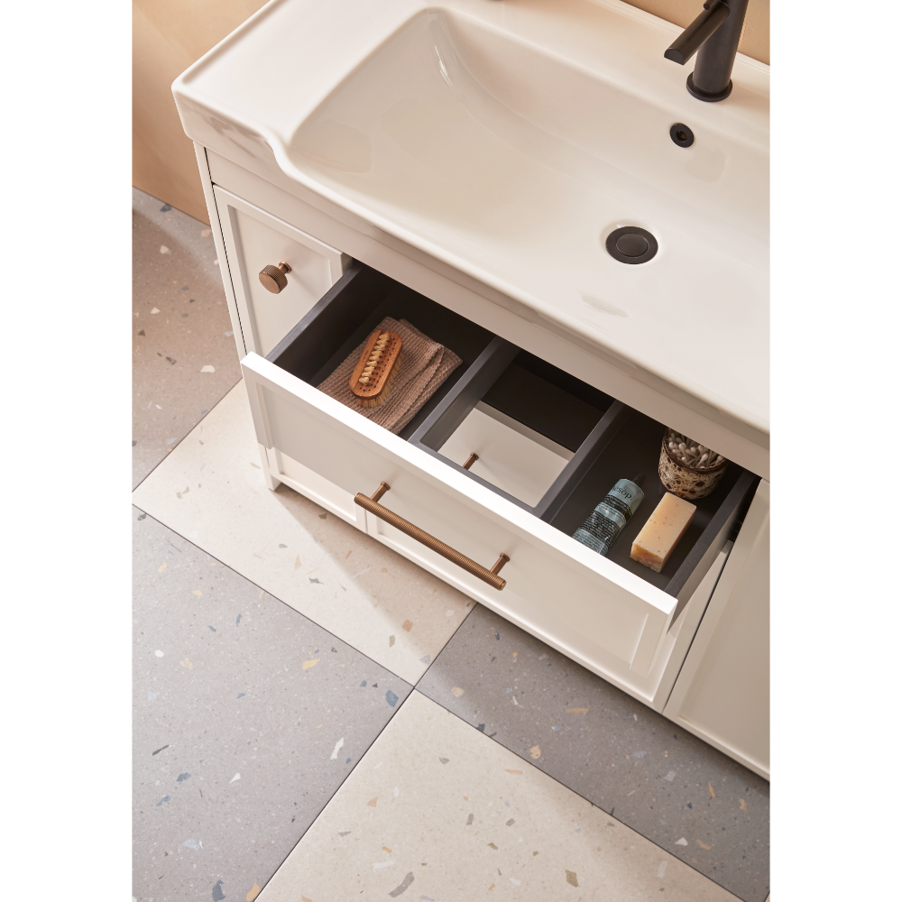 Roper Rhodes Concierge matt white vanity unit with white ceramic basin, bronze tap, and open drawer displaying organized bathroom storage with wooden brushes and toiletries, featuring brass handles and terrazzo flooring