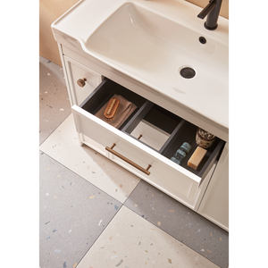 Roper Rhodes Concierge matt white vanity unit with white ceramic basin, bronze tap, and open drawer displaying organized bathroom storage with wooden brushes and toiletries, featuring brass handles and terrazzo flooring