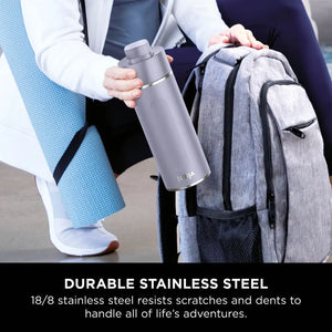 Person holding lavender Ninja Thirsti 700ml insulated stainless steel travel bottle next to grey backpack, showcasing durable 18/8 stainless steel construction