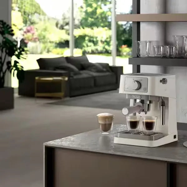 DeLonghi Stilosa cream espresso machine on grey kitchen counter with freshly brewed espresso cups, modern living room with black sofa and garden view in background