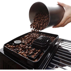 Hand pouring roasted coffee beans from a metal measuring cup into the bean hopper of a black DeLonghi Magnifica Start bean to cup coffee machine with control panel and chrome accents visible
