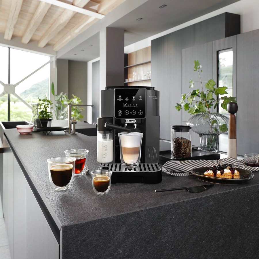 Delonghi Magnifica Start black bean to cup coffee machine on dark grey kitchen island countertop with espresso cups, coffee beans in glass container, pastries on plate, potted plants and window view in modern minimalist kitchen