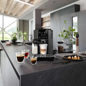 Delonghi Magnifica Start black bean to cup coffee machine on dark grey kitchen island countertop with espresso cups, coffee beans in glass container, pastries on plate, potted plants and window view in modern minimalist kitchen