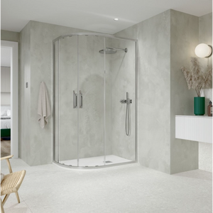 Modern chrome quadrant shower enclosure with curved glass doors in minimalist bathroom featuring light grey concrete-effect walls, white floor tiles, integrated rainfall showerhead and handheld spray, chrome fixtures, white vanity unit with green vase accent, and natural wood chair