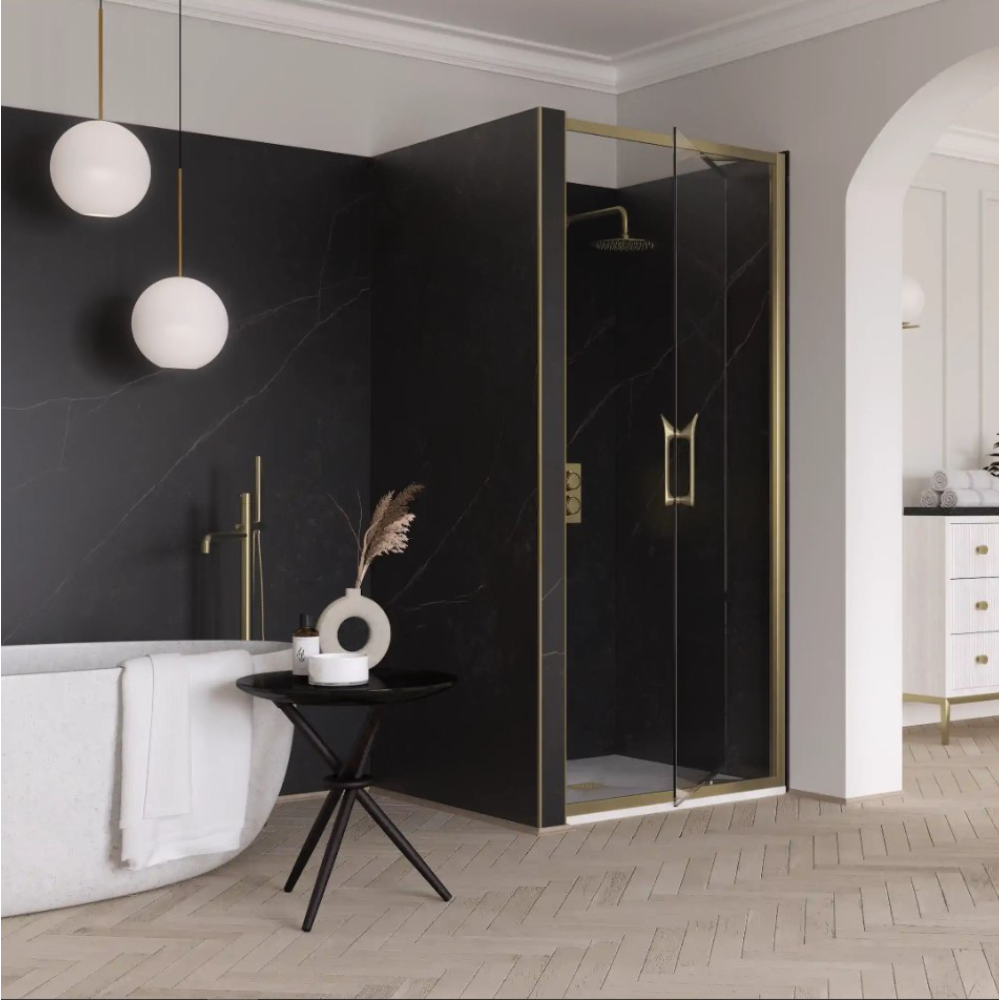 Modern luxury bathroom featuring Flair ETO Infold Shower Door with brushed brass frame and black marble panels, white freestanding bathtub, pendant sphere lighting, black tripod side table with white ceramic vase, white vanity with gold handles, and herringbone wood flooring