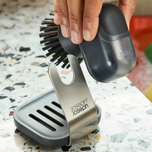 Hand holding black Joseph Joseph palm brush soap dispenser over stainless steel stand with bristles visible, displayed on speckled white countertop with cleaning debris
