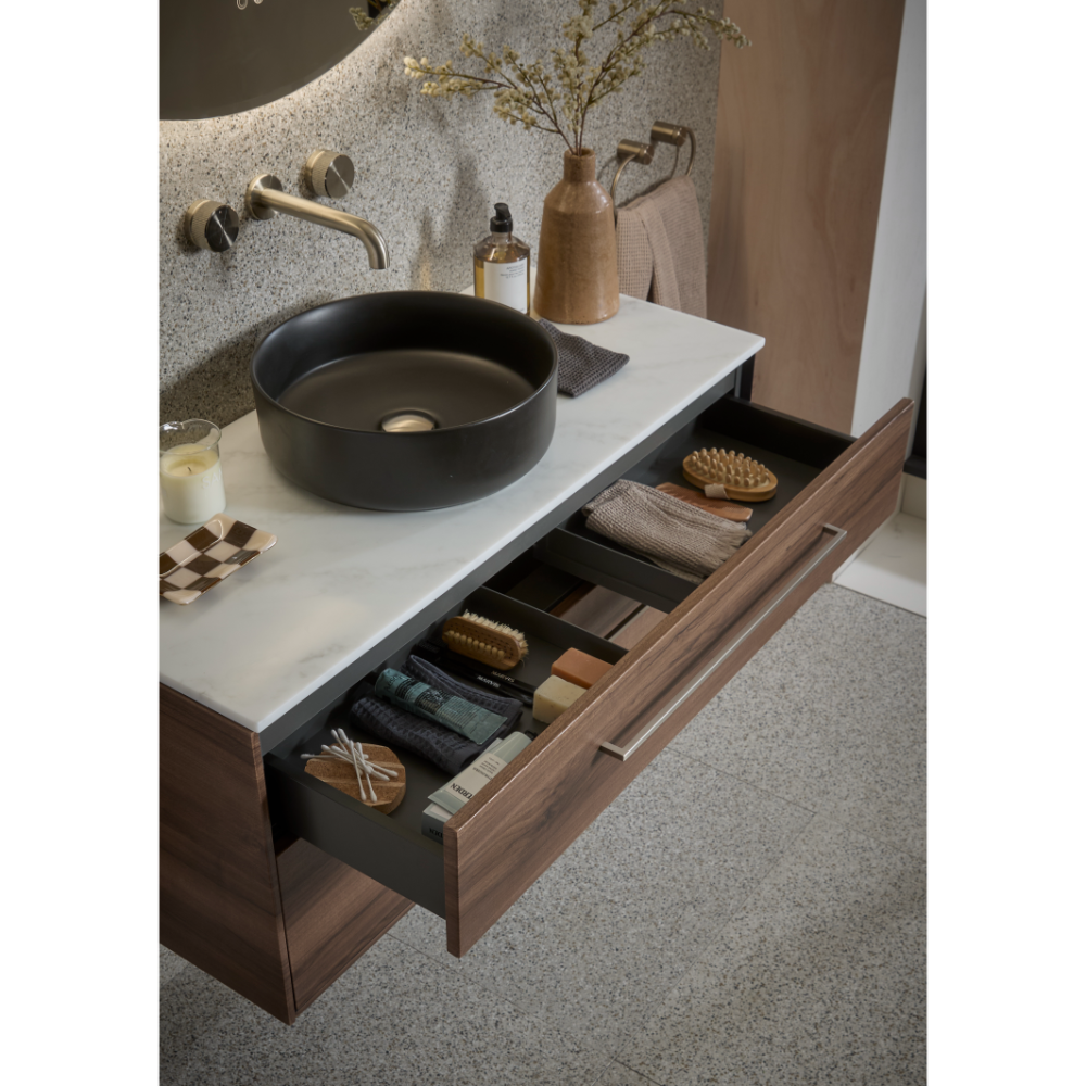 Modern bathroom vanity unit with Tuscan walnut wood base, white countertop and round black ceramic sink, featuring open drawer with organized storage including towels, brushes and bath accessories, brass wall-mounted faucet, textured stone backsplash, and decorative items including wooden vase with dried flowers and amber glass bottle