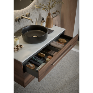 Modern bathroom vanity unit with Tuscan walnut wood base, white countertop and round black ceramic sink, featuring open drawer with organized storage including towels, brushes and bath accessories, brass wall-mounted faucet, textured stone backsplash, and decorative items including wooden vase with dried flowers and amber glass bottle