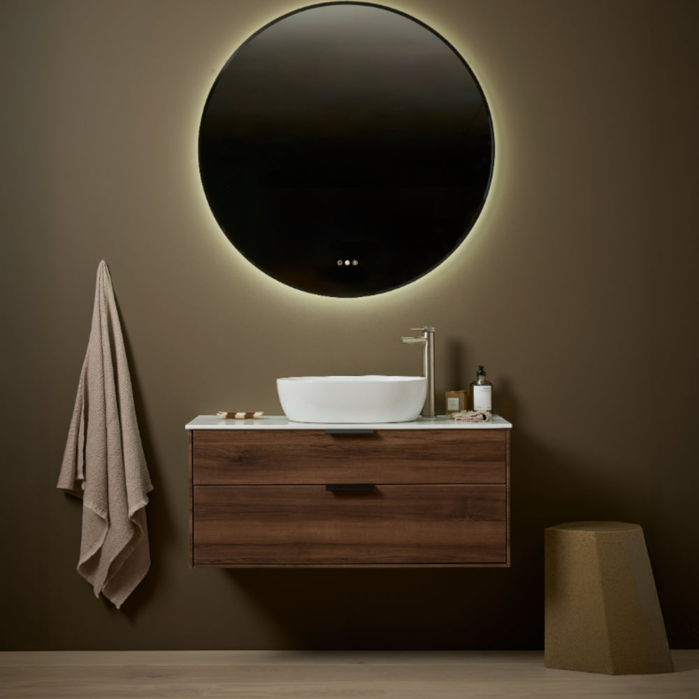 Modern bathroom vanity unit with Tuscan walnut finish featuring round backlit mirror, white vessel sink, chrome faucet, towel rail, and sage green accent stool against warm taupe wall