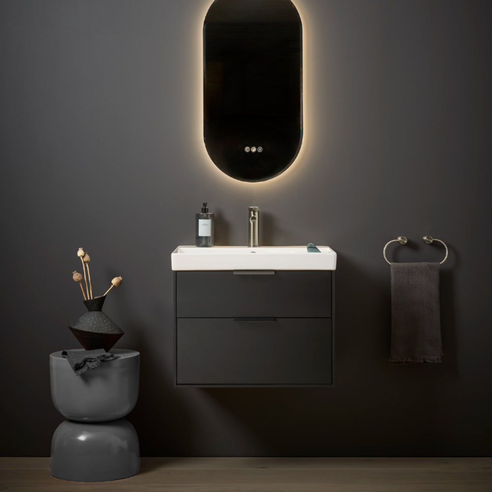 Modern black bathroom vanity unit with white ceramic sink, backlit oval mirror with warm ambient lighting, gold faucet, dark towel rail, and minimalist decor accessories on grey wall