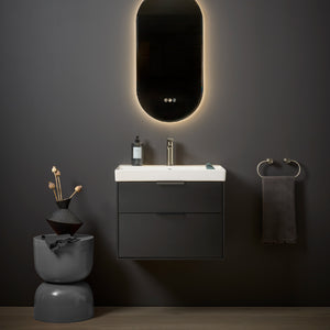 Modern black bathroom vanity unit with white ceramic sink, backlit oval mirror with warm ambient lighting, gold faucet, dark towel rail, and minimalist decor accessories on grey wall