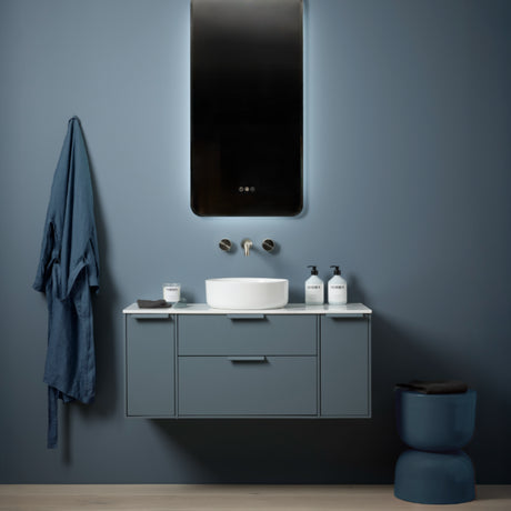 Modern bathroom vanity unit in sage green with white countertop, round white basin, chrome wall-mounted faucet, black framed mirror, and matching blue towel and waste bin accessories