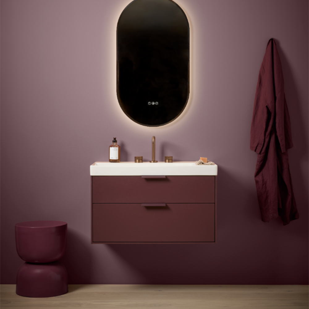 Modern burgundy bathroom with Roper Rhodes Faber wall-mounted vanity unit featuring white countertop and burgundy cabinetry, oval backlit mirror with warm glow, brass fixtures, matching burgundy stool and towels, wooden floor, contemporary minimalist design